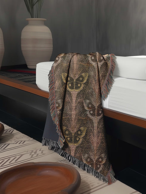 A woven cotton throw featuring an intricate luna moth pattern in bronze and muted earth tones, framed by soft fringe.